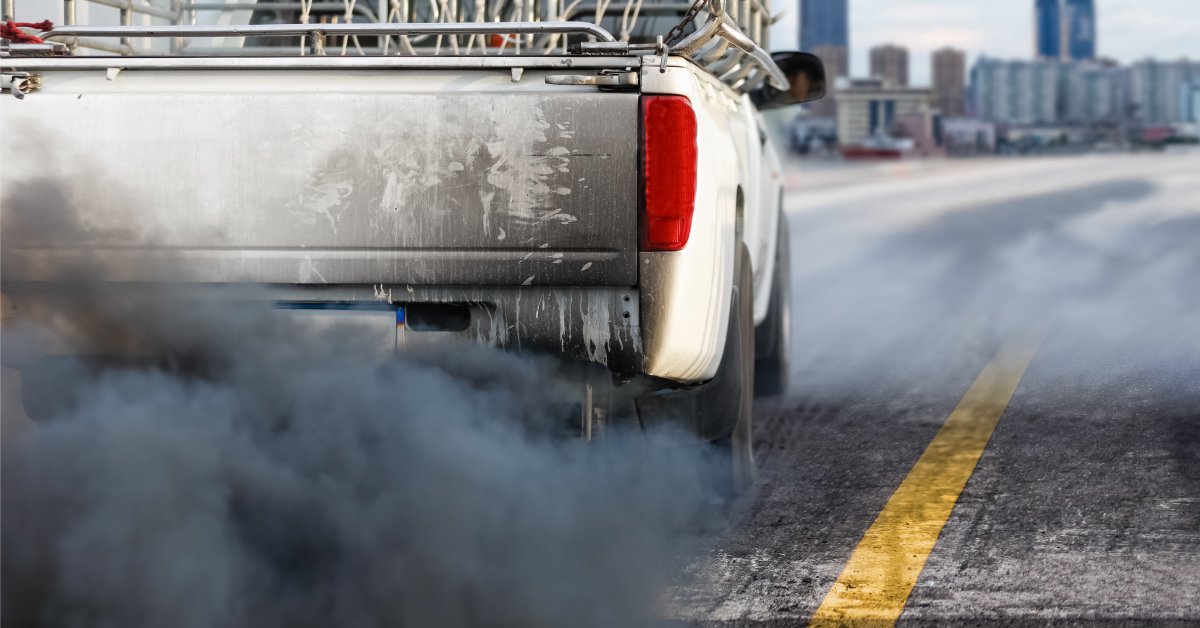 A truck drives down a highway. Dark smoke is coming out of the exhaust pipe, filling the air behind the truck.