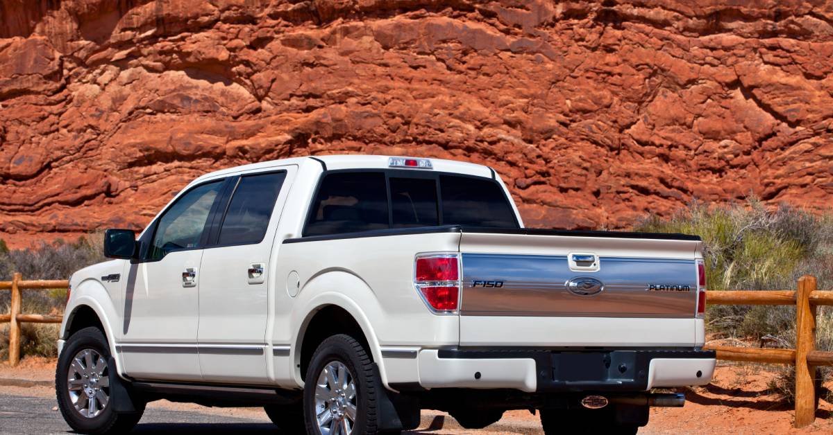 A white pickup truck drives down the road with a fence to its right, some greenery, and a large red rock formation.